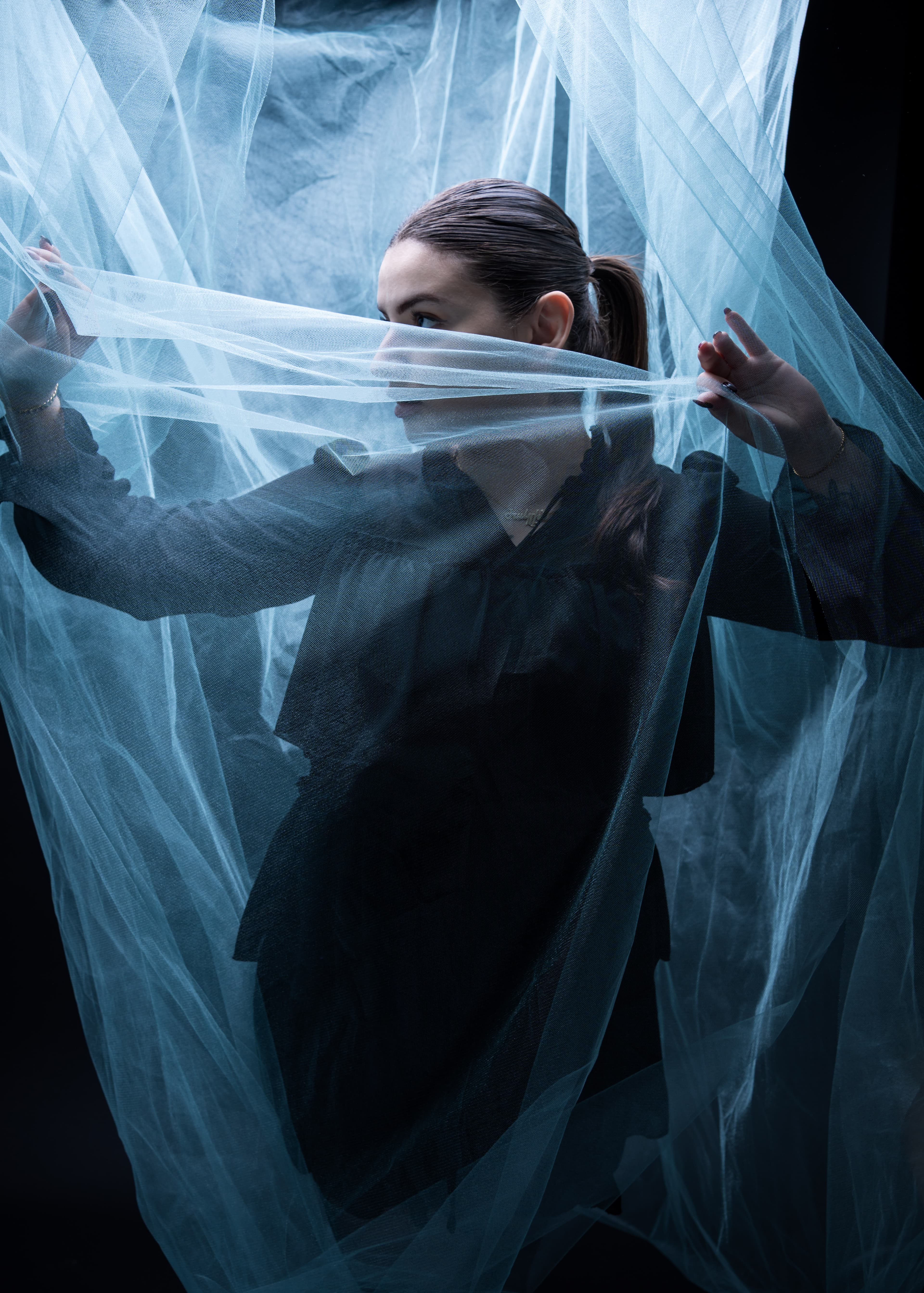 Frau in dunkler Kleidung zwischen blauem Tüllstoff, seitliche Pose im Studio